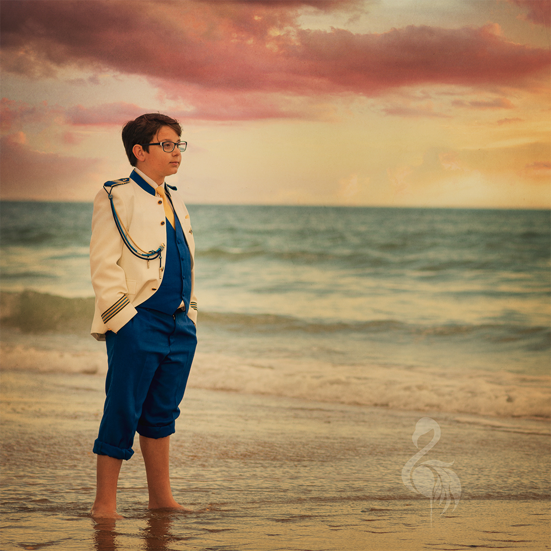 Niño vestido de traje de comunión en la orilla de la playa al atardecer, mirando hacia el horizonte con el mar de fondo