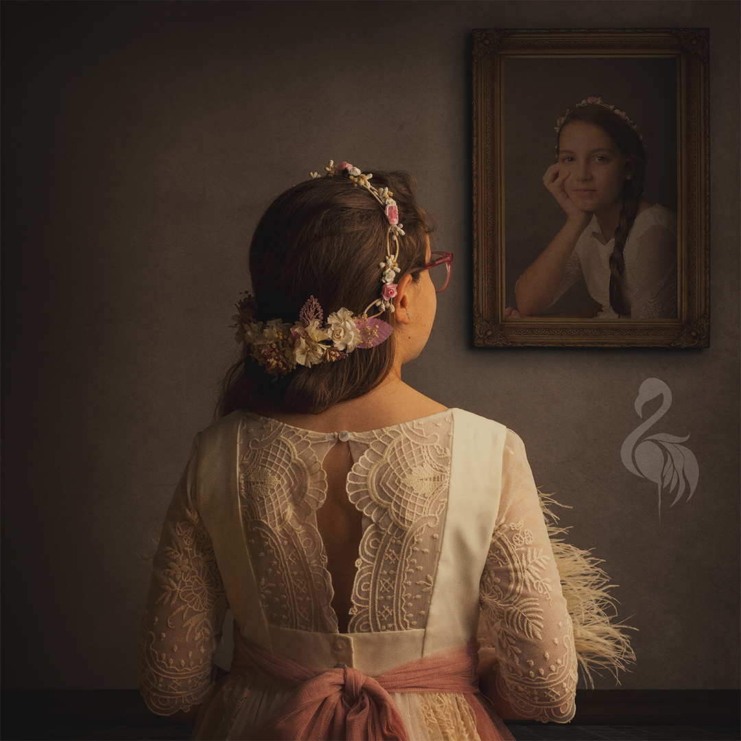 Niña con vestido de comunión de encaje, vista de espaldas mientras mira un retrato antiguo de sí misma en la pared